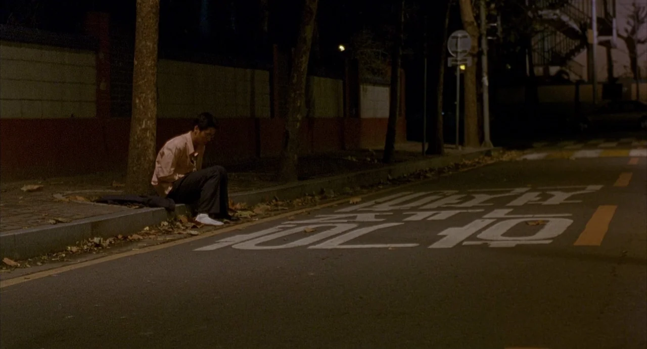Barking Dogs Never Bite (2000) movie still: wide — A dark urban street at night with white Korean traffic markings painted on asphalt. A lone man…