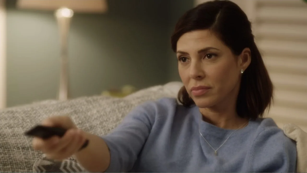 A Christmas Break (2020) movie still: medium — A domestic living room space features a grey textured sofa and a lamp. A woman in a blue sweater sits…