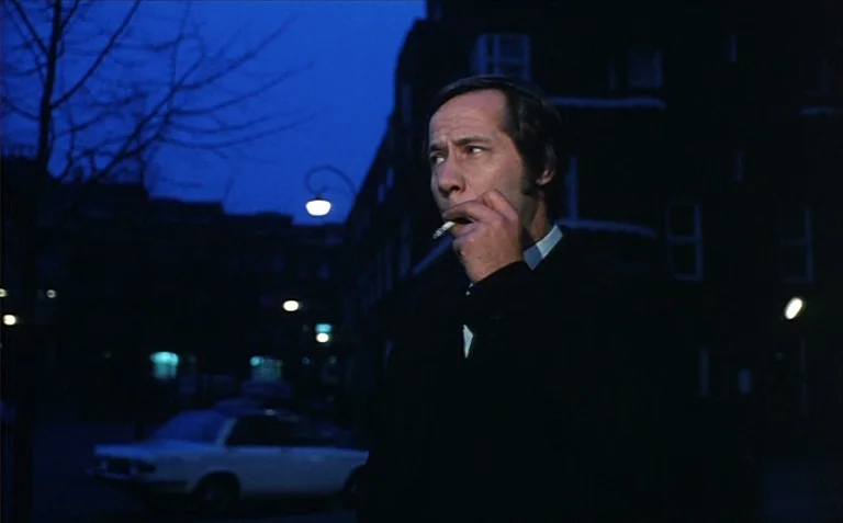 Business is Business (1971) movie still: medium — A dark city street at night features buildings in silhouette and a parked car. A middle-aged man…