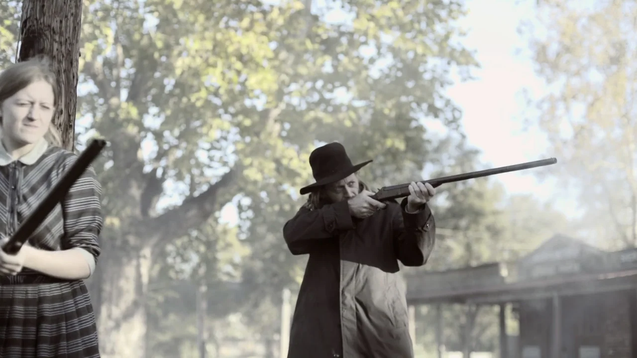 The Dalton Gang (2020) movie still: medium — An outdoor frontier setting features trees and wooden structures in the soft daylight. A man with a…