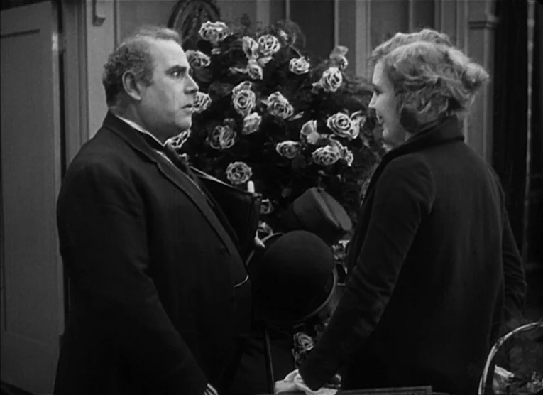 The Kid (1921) movie still: medium — An interior parlor features a large, dense floral arrangement centered between two people. A man in a dark suit…