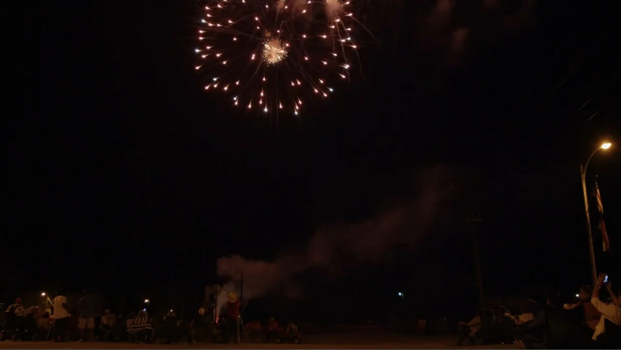 Rich Hill (2014) movie still: wide — A nighttime outdoor setting with a small crowd sitting in chairs during a firework show. Large white and gold…