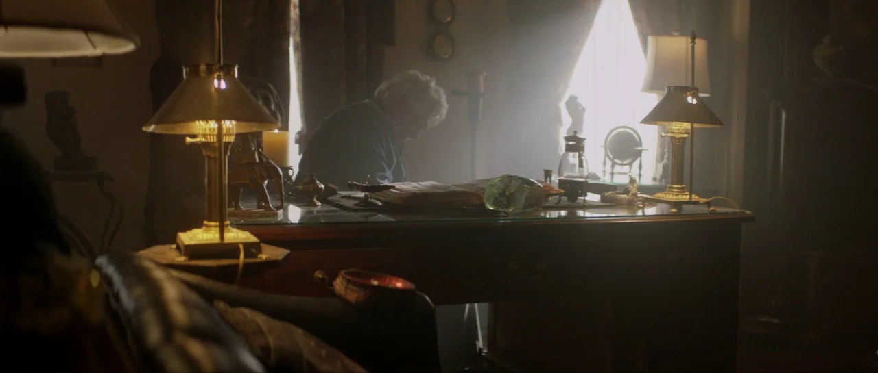 Suitable Flesh (2023) movie still: wide — A dimly lit study contains a large wooden desk filled with various ornamental items. An older man sits at…