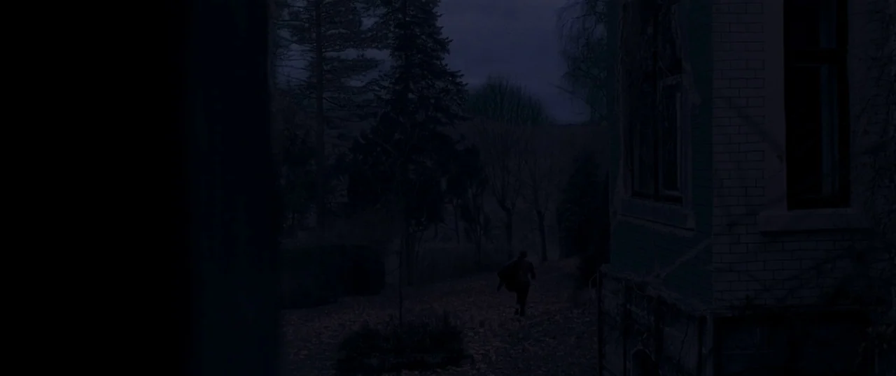 Personal Shopper (2016) movie still: wide — A dark residential yard at night contains a brick building corner and sparse trees. A lone figure runs…