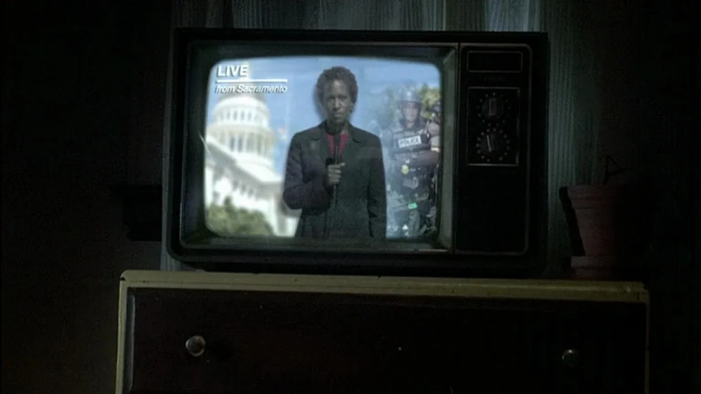 Land of Plenty (2004) movie still: medium — An old cathode ray tube television sits on a dark wooden chest in a room. A news broadcast showing a…