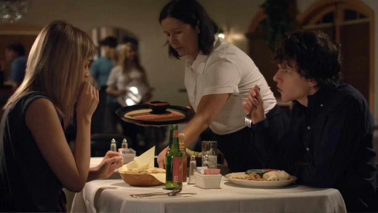 Free Samples (2013) movie still: medium — A restaurant interior features a white-clothed dining table holding plates and a glass bottle. A waitress…