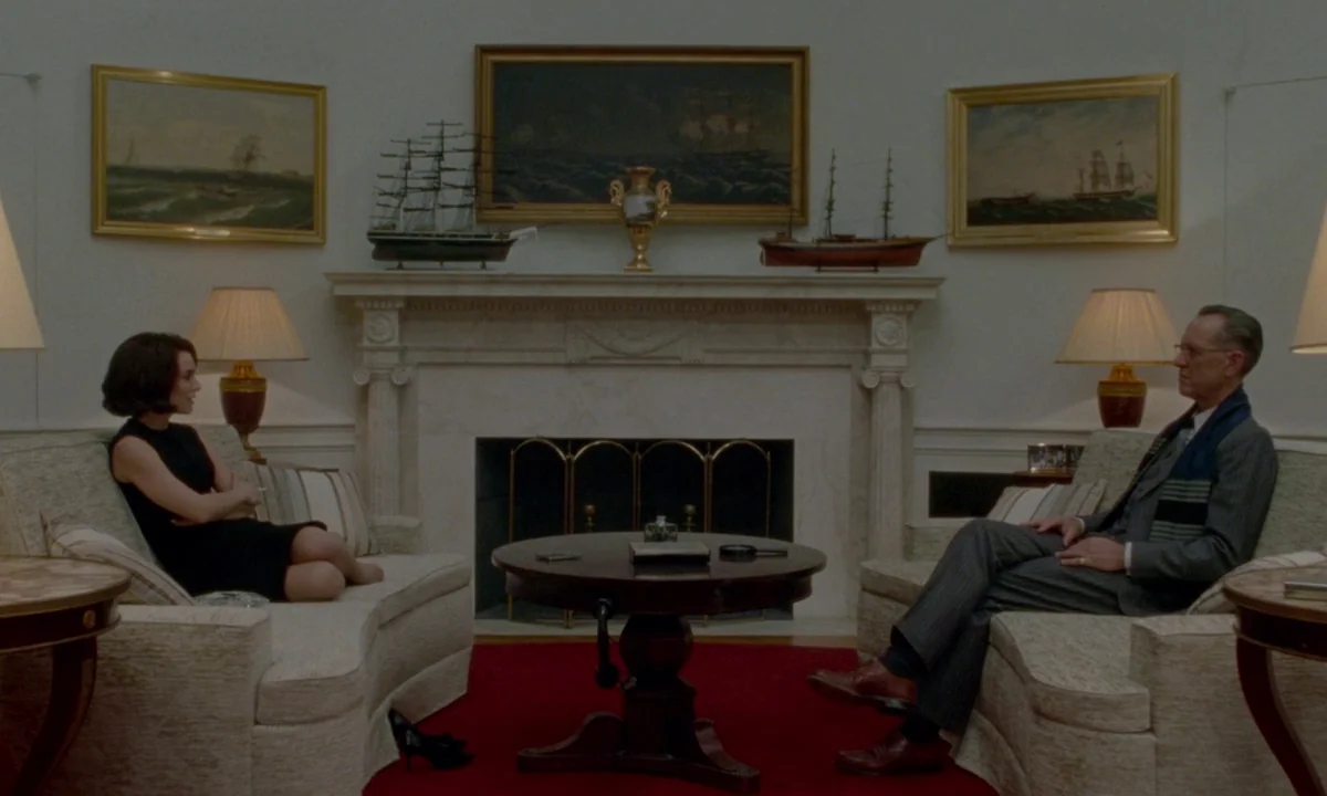 Jackie (2016) movie still: wide — An upscale room featuring a marble fireplace flanked by matching sofas and ship models. A woman and a man sit…