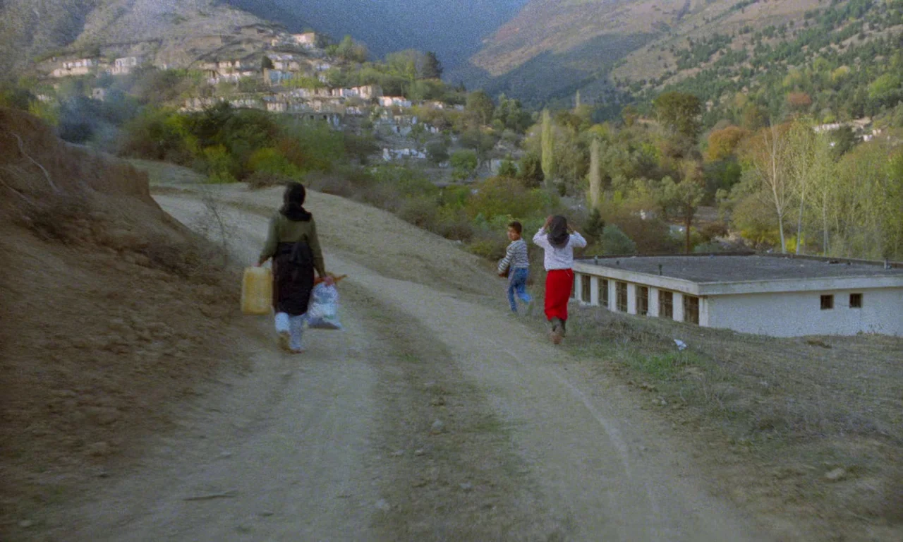 Life, and Nothing More (1992) movie still: wide — A dusty dirt path winds through a mountainous rural area near a small village. Three people walk…