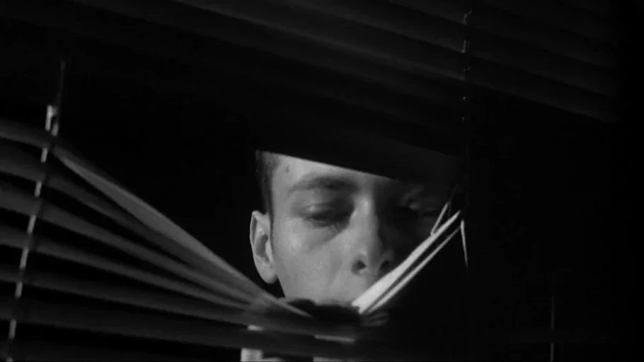 American History X movie still: closeup — A dark room is punctuated by light filtering through horizontal venetian blinds. A man peers through the…