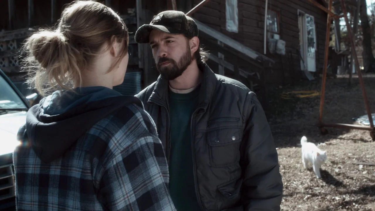 Winter’s Bone (2010) movie still: medium — A man and woman talk outdoors near a wooden structure and a parked vehicle. A white cat walks across the…