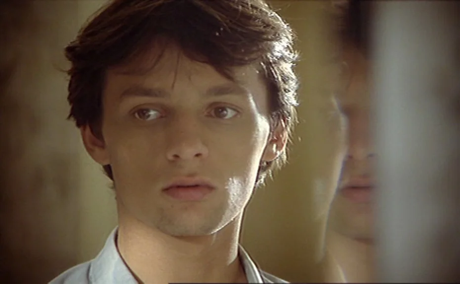 Diva (1981) movie still: closeup — A young man gazes into a mirror inside a dimly lit domestic bedroom space. The reflection of his face is clearly…