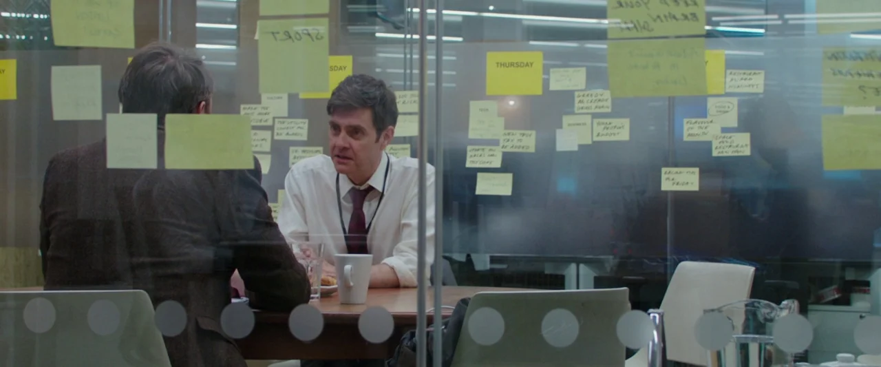 Greed (2019) movie still: medium — A glass-walled meeting room features multiple yellow sticky notes attached to the transparent surface. Two men…