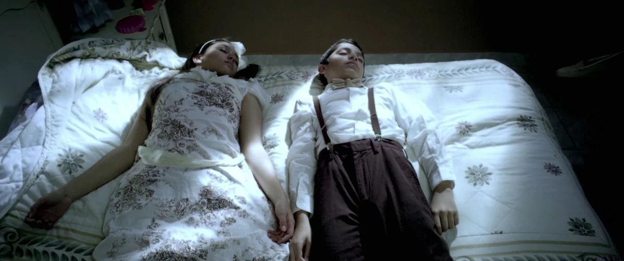 Here Comes The Devil (2012) movie still: medium — Two adolescents lie flat on their backs across a bed with a floral quilt. The pair are dressed in…