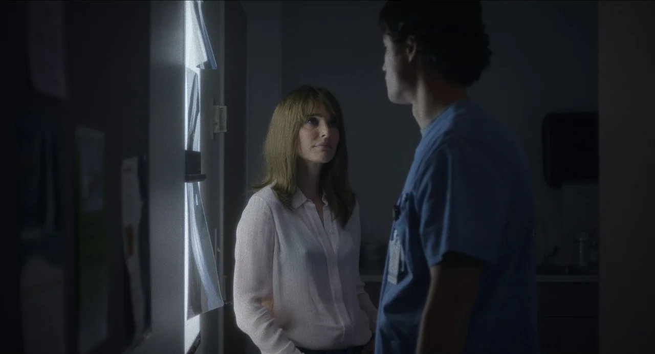 May December (2023) movie still: medium — A dimly lit room features a medical X-ray viewer mounted on a wall. A woman in a light shirt speaks to a…
