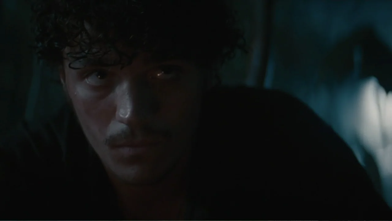 Jurassic World Rebirth (2025) movie still: closeup — A dark, dimly lit indoor space with indistinct shadows forming the background. A young man with…