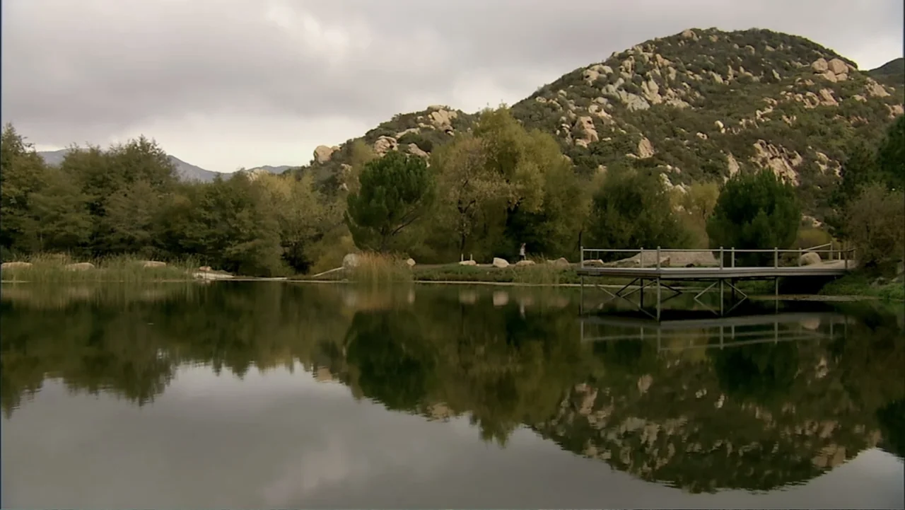 Hot Gimmick: Girl Meets Boy (2019) movie still: establishing — A tranquil pond sits at the base of a rocky, tree-covered hillside under overcast…