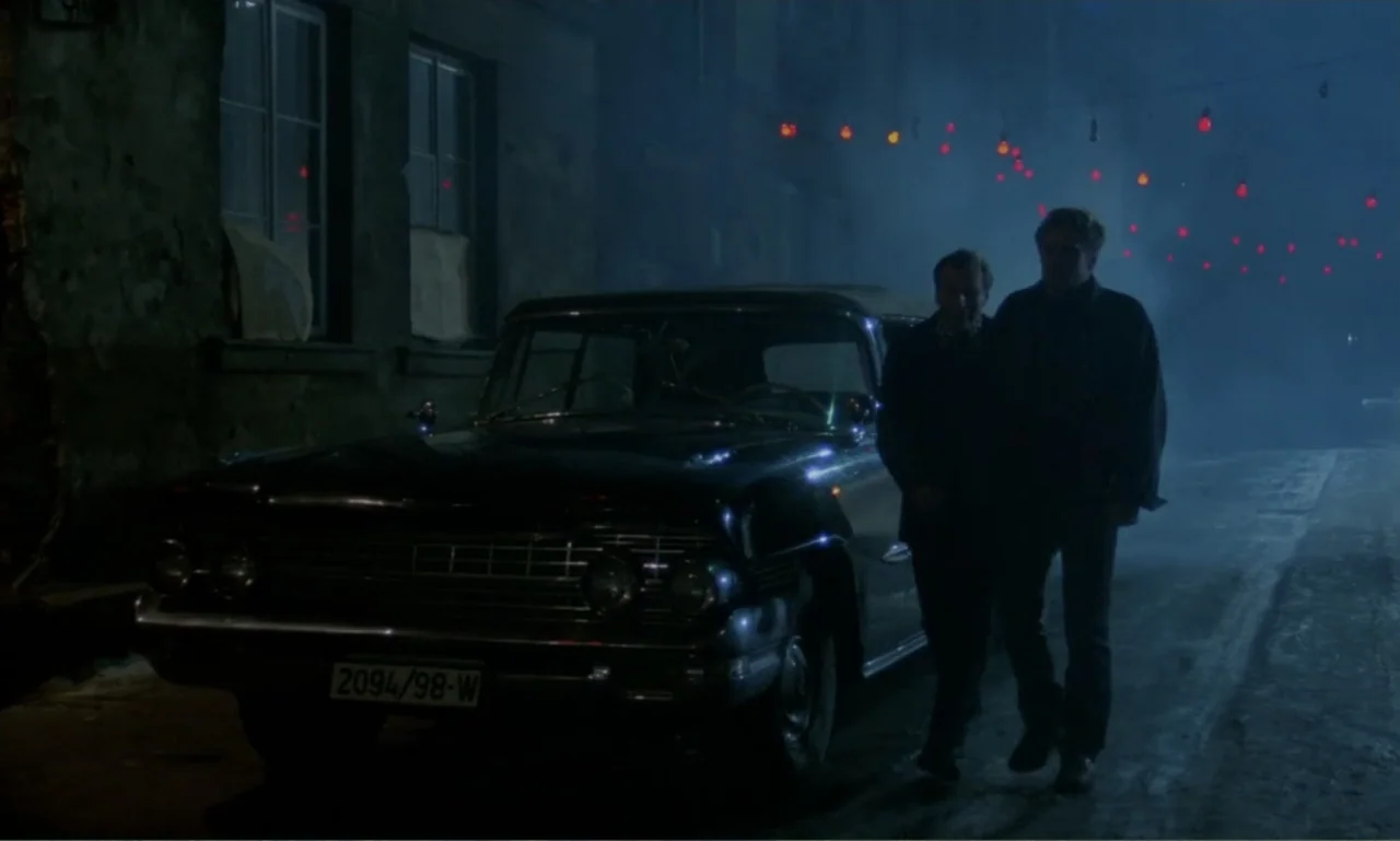 Ga-ga: Glory to the Heroes (1986) movie still: wide — A dark night scene shows a vintage black sedan parked along an old building wall. Two men walk…