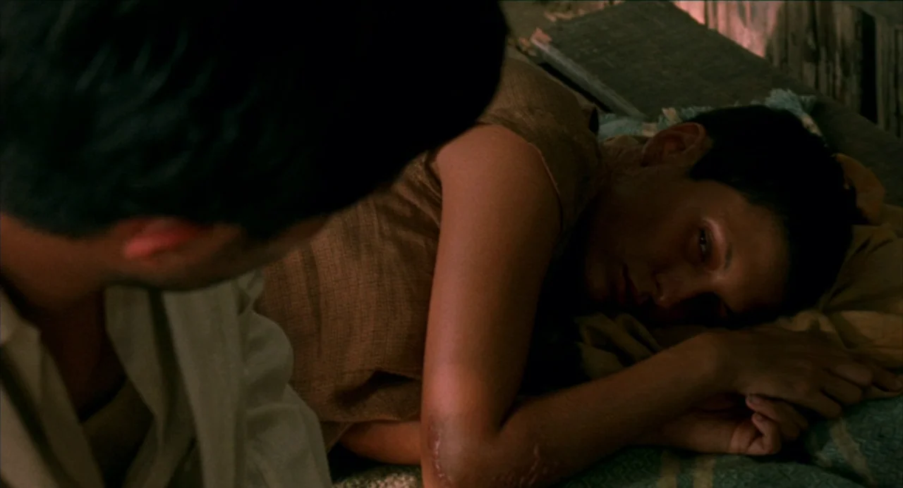 The Motorcycle Diaries (2004) movie still: medium — A dimly lit room with simple furnishings. A woman lies on a bed while a man sits nearby.