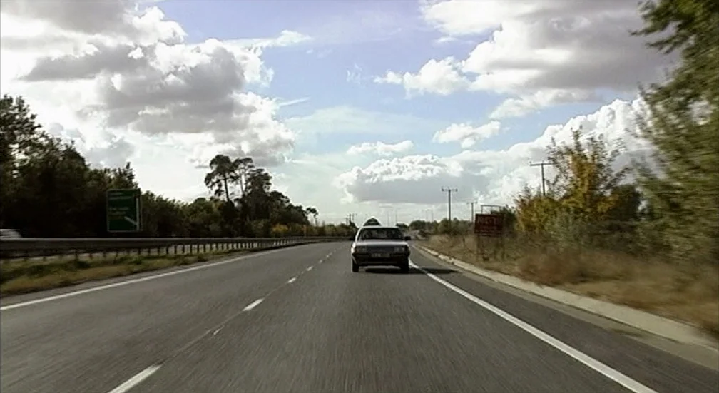 9 Songs (2004) movie still: wide — A wide multi-lane highway stretches toward the horizon under a cloudy sky. A silver sedan travels along the road…