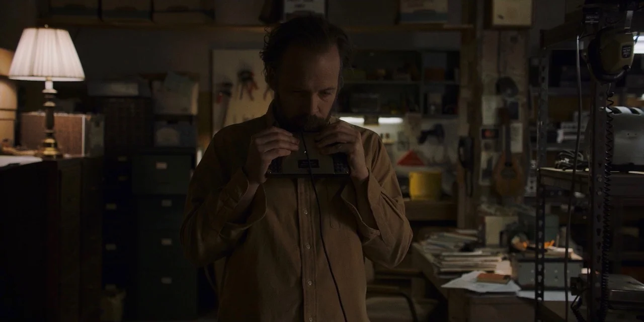 The Sound of Silence movie still: medium — A dark, cluttered workspace is filled with electronic equipment, shelves, and various loose items. A man…
