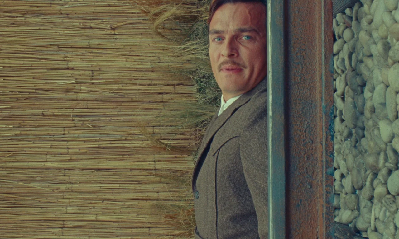 The Swan (2023) movie still: medium — A man stands outdoors beside a vertical split between a woven reed screen and stone wall. The man is…