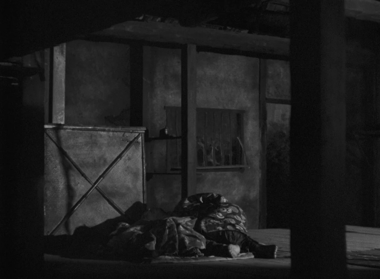 Ugetsu (1953) movie still: wide — A dimly lit room with rough walls and a wooden structure in the background. A person sleeps under a patterned…