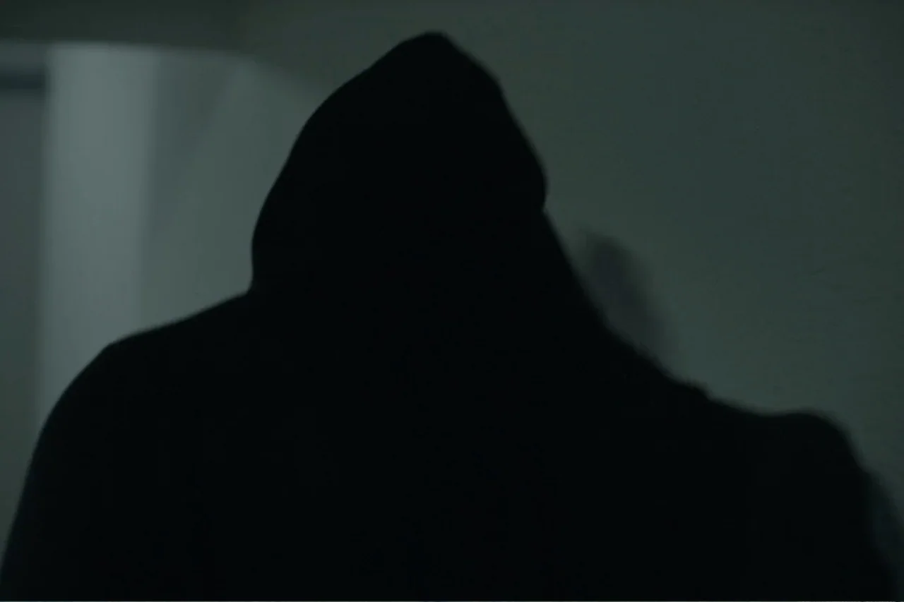Cross the Line (2020) movie still: medium — A dark, featureless interior wall provides a backdrop for a solitary hooded figure. The subject faces…