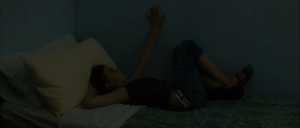 Genova (2008) movie still: medium — A dimly lit room with blue walls and a small bed against the wall. A young person lies on their side with an arm…