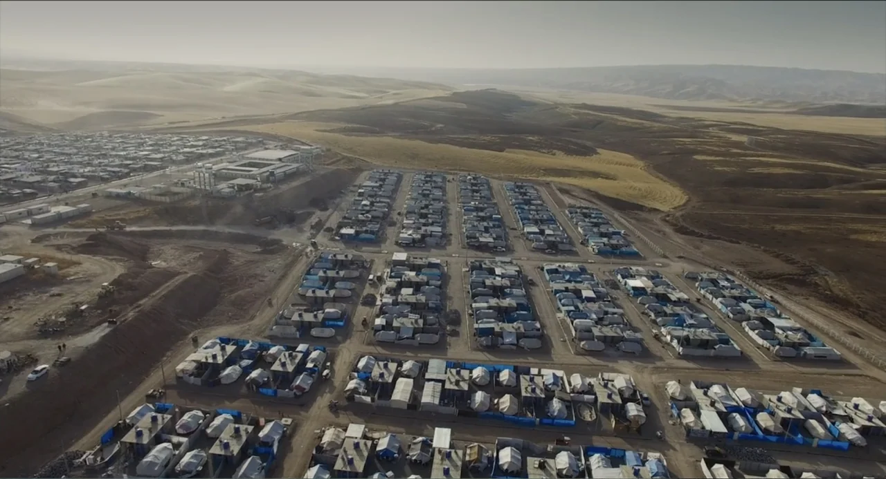Human Flow (2017) movie still: establishing — A sprawling encampment of temporary shelters sits on a dry, arid plain. Rows of tents are arranged in…