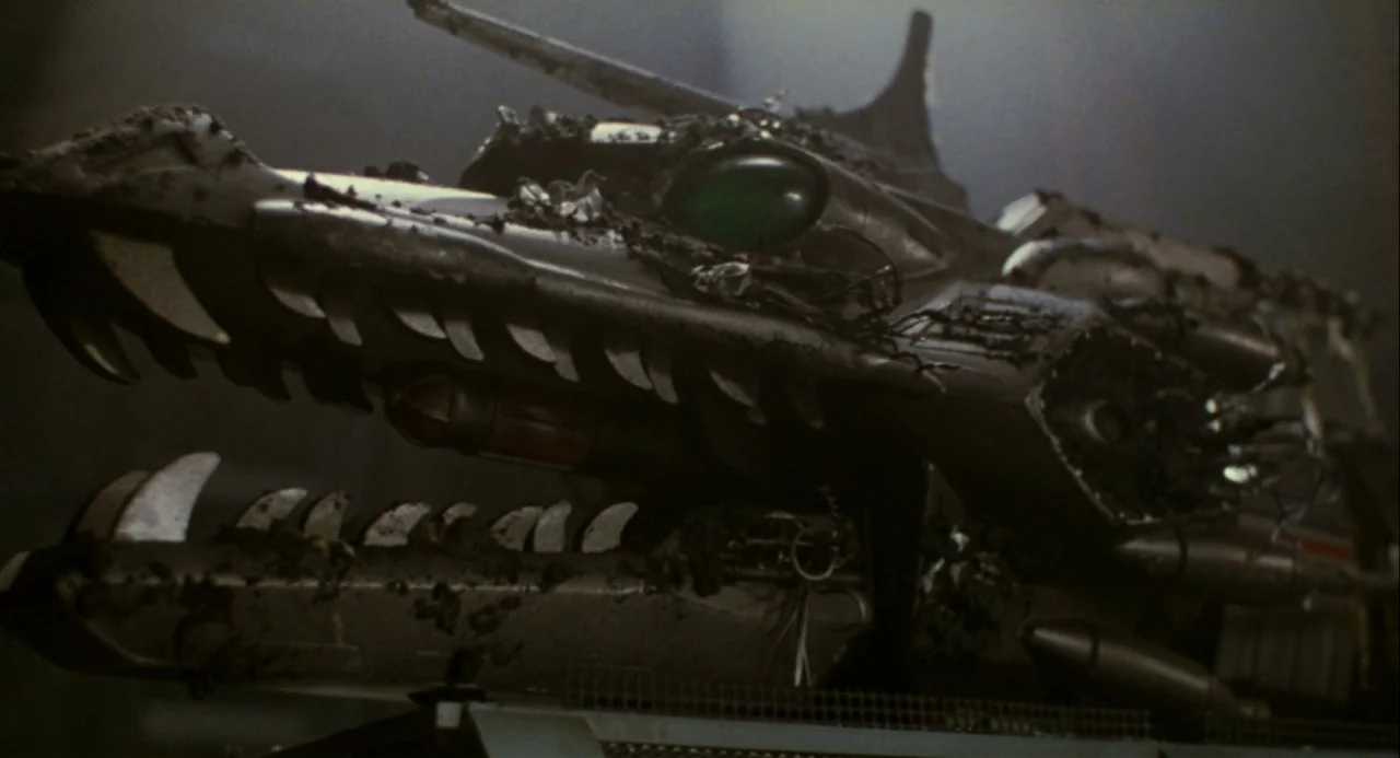Godzilla vs. Mechagodzilla II (1993) movie still: closeup — A detailed mechanical dragon head sits within a dimly lit industrial space. Debris and…