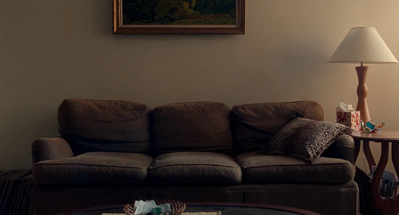 His Three Daughters (2023) movie still: wide — A brown fabric sofa sits centered against a plain beige wall under a landscape painting. A wooden end…