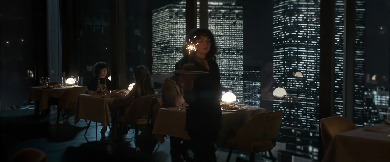 Drop (2025) movie still: medium — A dark restaurant interior at night with large windows overlooking a city skyline. A woman stands holding a…