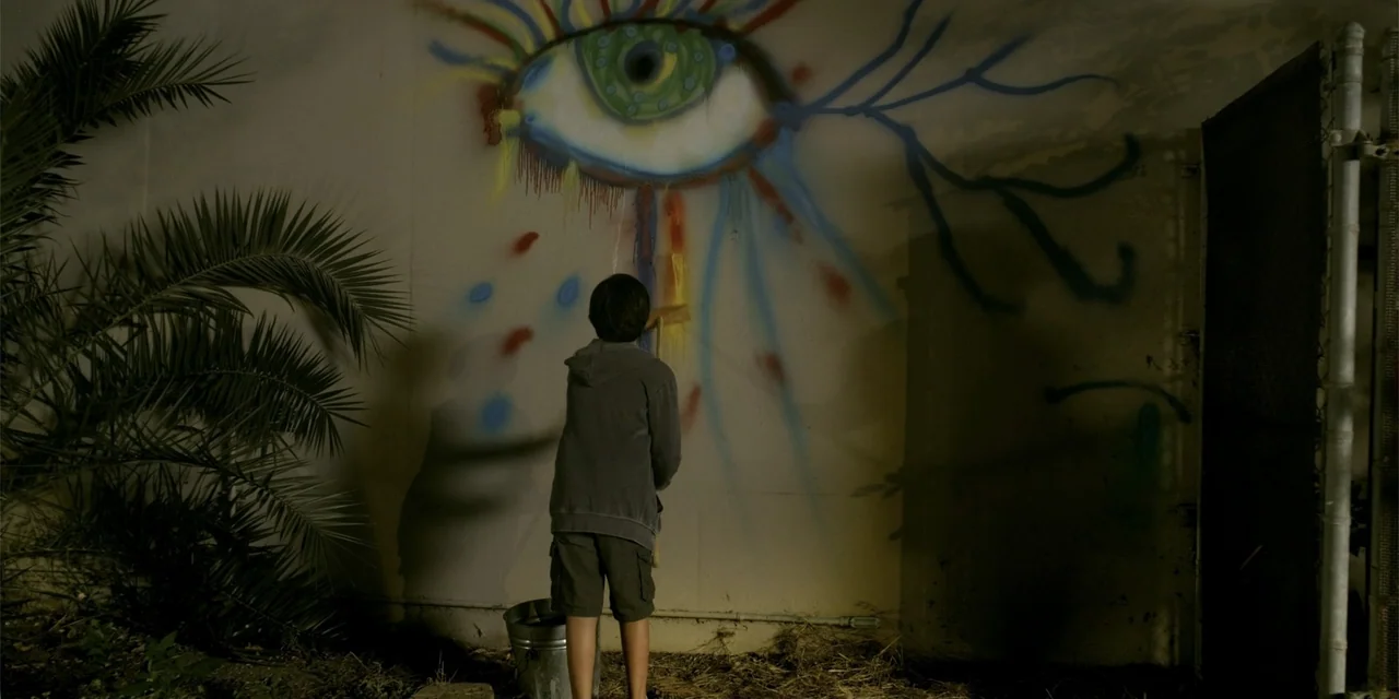 7th & Union (2021) movie still: fullbody — A concrete wall in an alley serves as a canvas for a large eye mural. A young child wearing a hoodie…