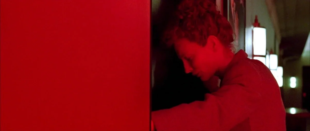 The Aviator (2004) movie still: medium — A dimly lit indoor passage is saturated in deep red light. A woman stands with her face pressed against a…