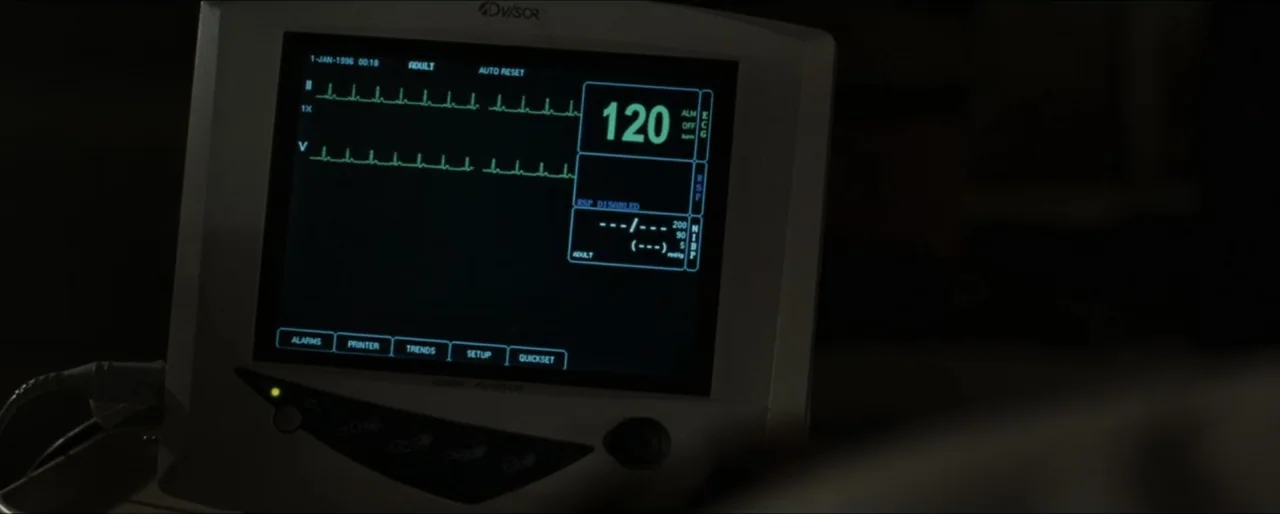Law Abiding Citizen (2009) movie still: closeup — A medical monitor sits in a dimly lit space displaying a heart rate reading. Green digital text…