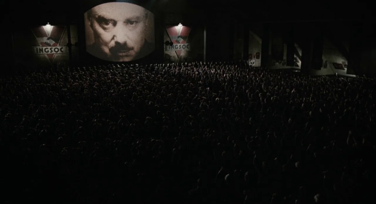1984 (1984) movie still: wide — A dark, cavernous interior filled with a dense crowd facing a large projection screen. Large posters featuring red…