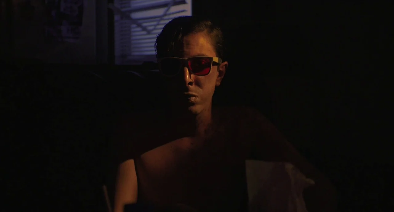 Relaxer (2018) movie still: medium — A man sits shirtless in a dark, shadow-filled room illuminated only by screen light. He wears vintage red and…