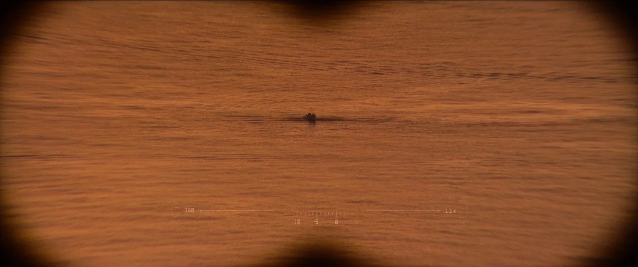 Pacifiction (2022) movie still: wide — An expanse of calm water at sunset is viewed through a circular optical device. A small, distant boat is…