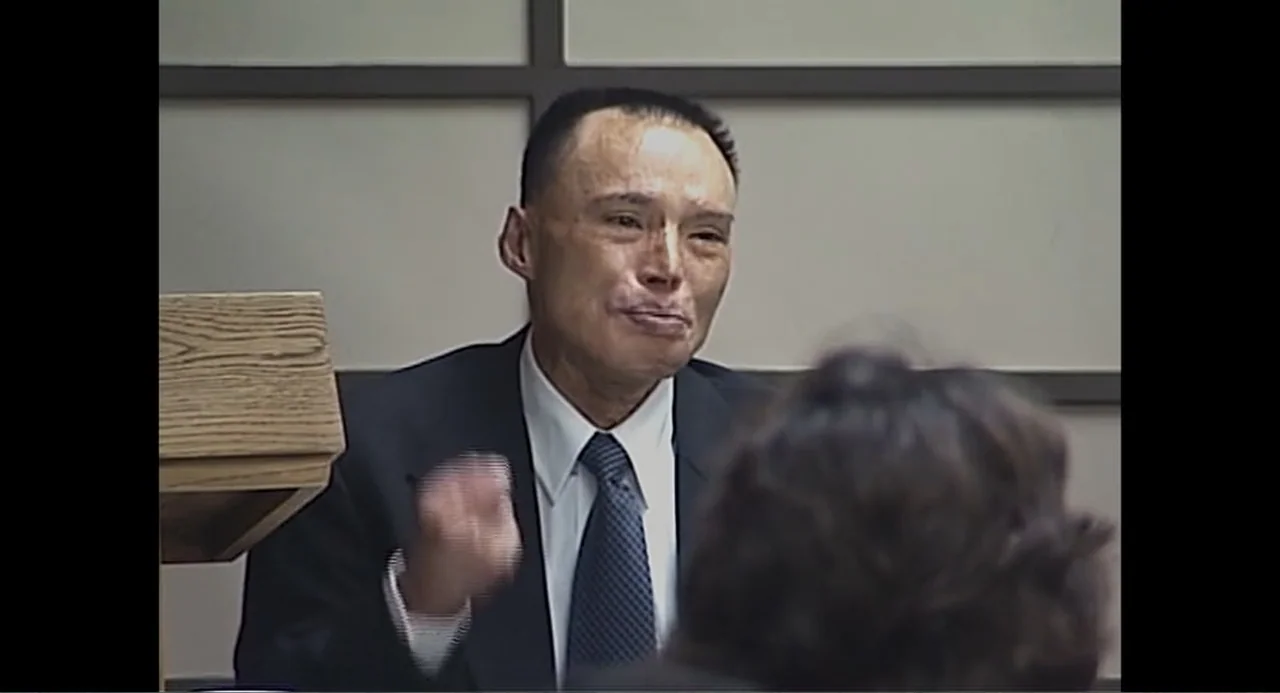Free Chol Soo Lee (2022) movie still: medium — A deposition room with an off-white grid-paneled wall and a wooden podium. A middle-aged man in a…