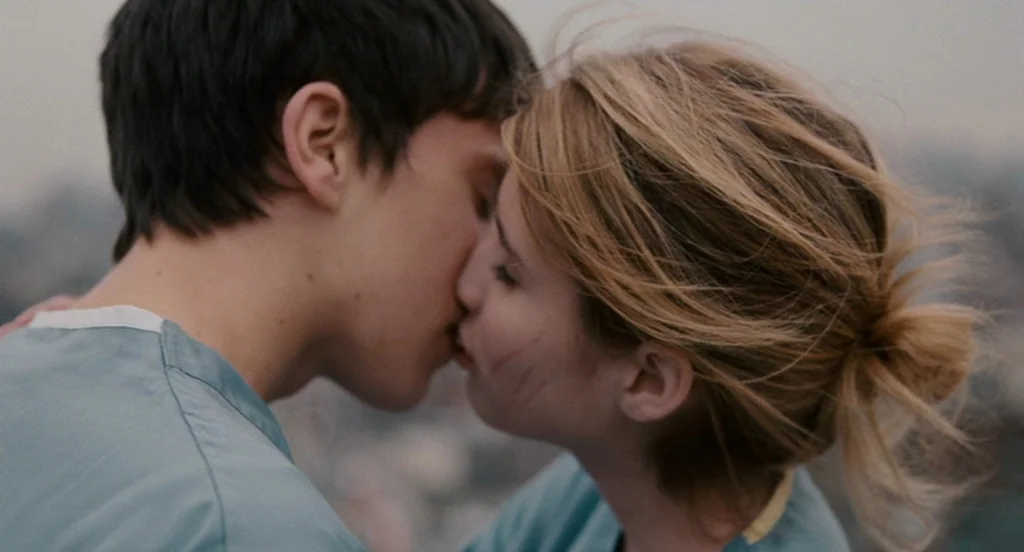 It’s Kind Of A Funny Story (2010) movie still: closeup — Two individuals wearing teal scrub tops embrace while kissing on a city rooftop. The soft…