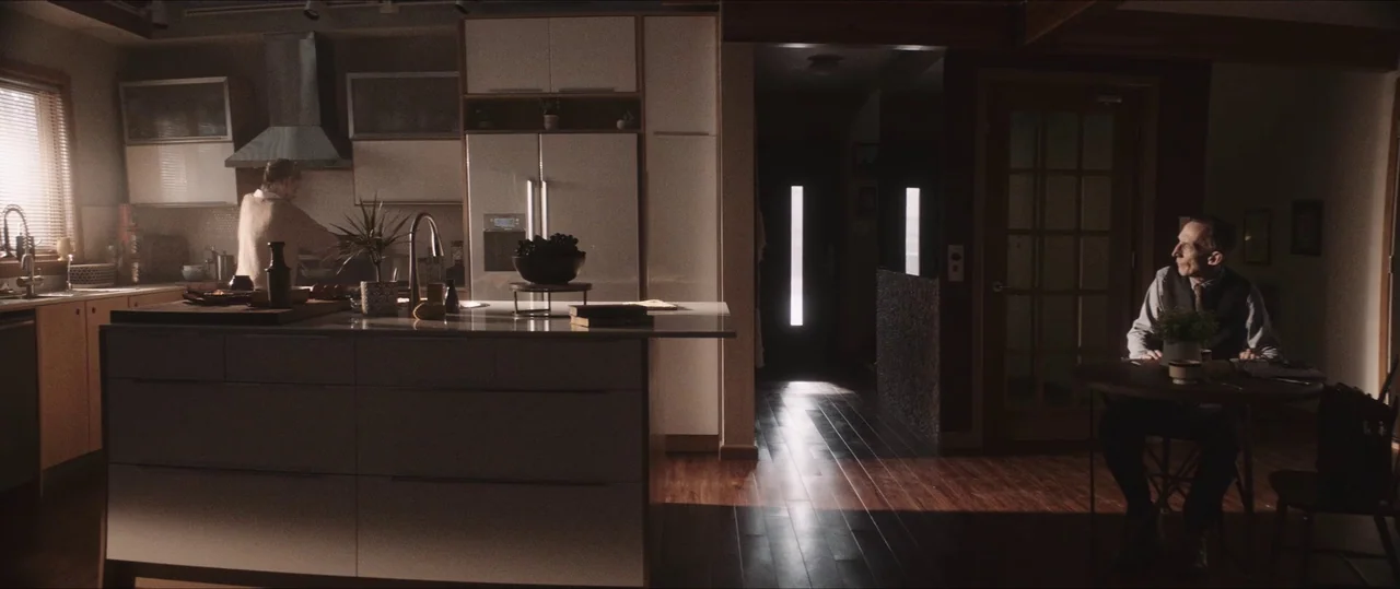 Anything For Jackson (2020) movie still: wide — A spacious home interior features a modern white kitchen island and dark wooden flooring. An elderly…
