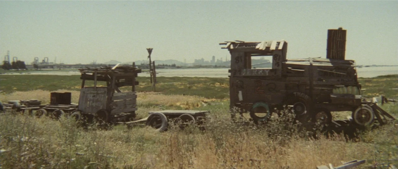 Take Me Away! (1978) movie still: wide — A makeshift wooden locomotive structure sits in a dry, grass-covered field near water. Distant city…