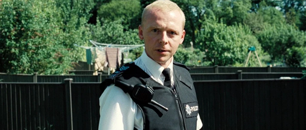 Hot Fuzz (2007) movie still: medium — A man in a police uniform stands in a residential backyard with wooden fencing. Behind him are green trees and…