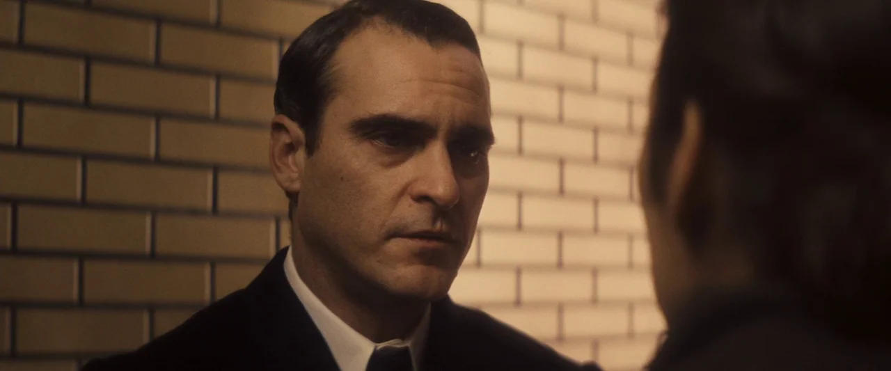The Immigrant (2013) movie still: closeup — An interior space features light brown brick walls arranged in a grid pattern. A man in a dark suit…