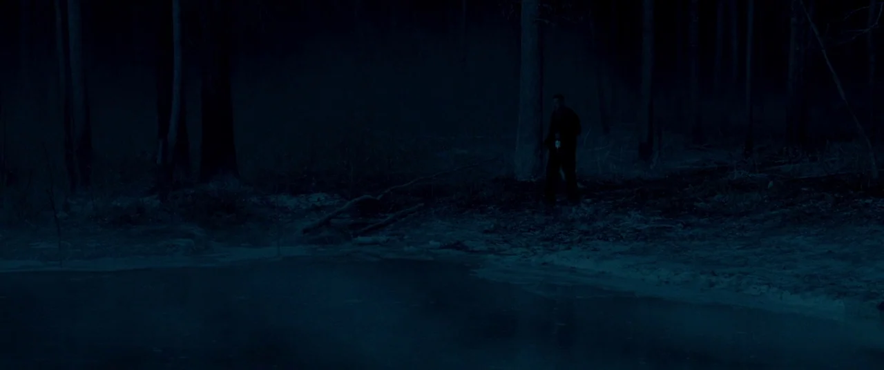 Lawless (2012) movie still: wide — A dark, wooded shoreline borders a calm pond under the dim light of night. A lone man in a dark coat stands near…