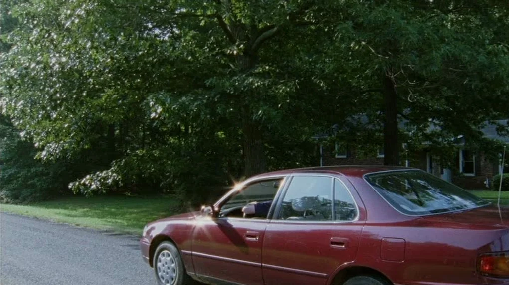 Junebug (2005) movie still: wide — A maroon sedan sits on the shoulder of a quiet road lined with mature trees. A driver operates the vehicle while…