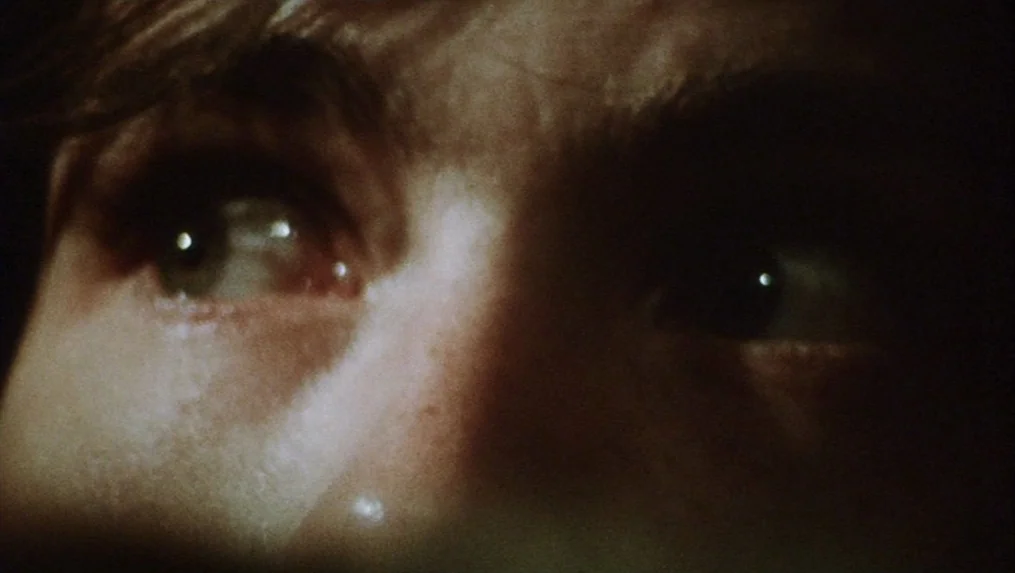 Martin (1977) movie still: closeup — A dark, obscured environment surrounds a close-up of a human face in shadow. The subject stares directly…