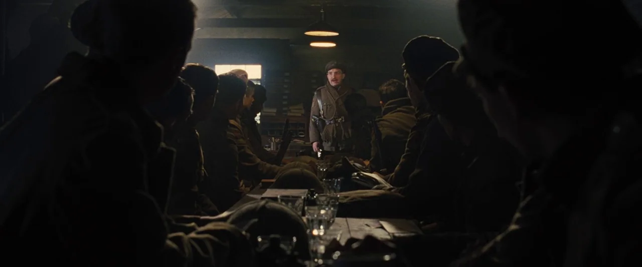 War Horse (2011) movie still: wide — A dimly lit room serves as a military barracks with long wooden tables and chairs. A soldier stands at the head…