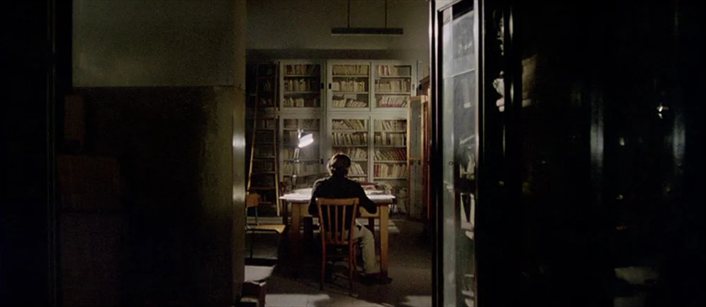 Deep Red (1975) movie still: wide — A dimly lit room features tall glass-fronted shelves packed with numerous books. A person sits at a wooden table…