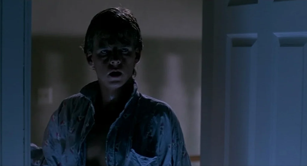 A Nightmare On Elm Street 2: Freddy’s Revenge (1985) movie still: medium — A dark, dimly lit indoor space frames a teenager standing in a doorway.…
